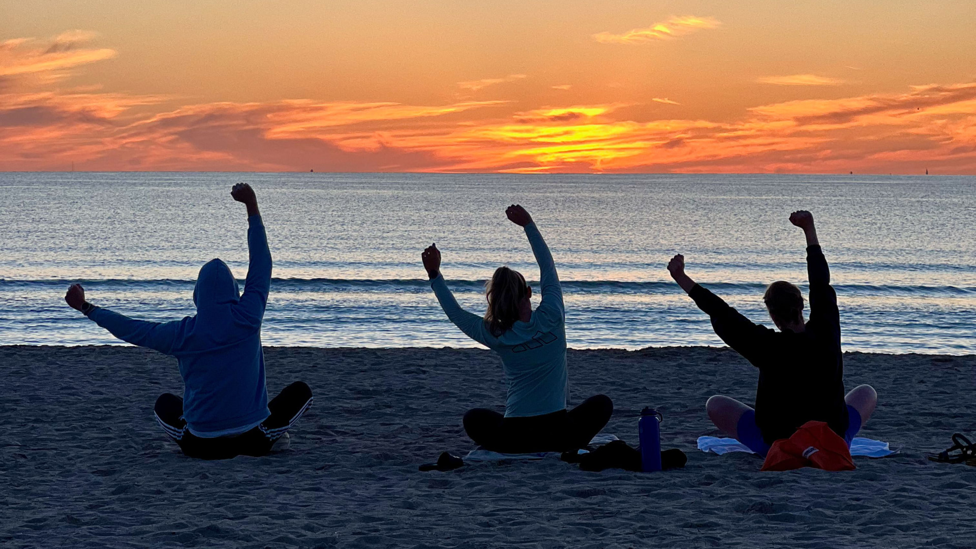 Three people stretching on the sand facing the ocean at sunrise